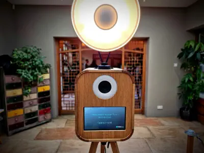 A rectangular wooden photobooth stands indoors on a tripod, with a circular ring light attached to the top. Behind it, there is a wooden door with a geometric pattern, and plants and drawers are visible on either side. A screen displays photo booth hours.