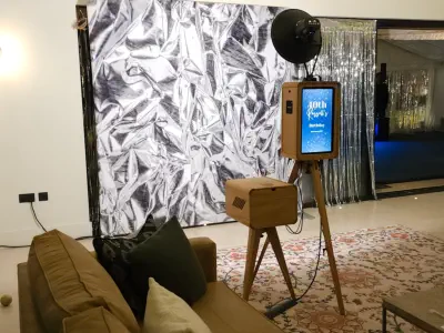 A modern Oxfordshire photo booth setup with a wooden frame and camera on a tripod stands in front of a silver, reflective backdrop. A couch with cushions is in the foreground on an ornate rug. The backdrop is in a spacious room with a shiny curtain to the