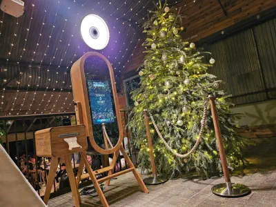 A festive setting features a decorated Christmas tree adorned with lights and ornaments. Nearby, a wooden photo booth hire Wiltshire with a touch screen sits under a ceiling covered in twinkling fairy lights, creating a warm and celebratory atmosphere.