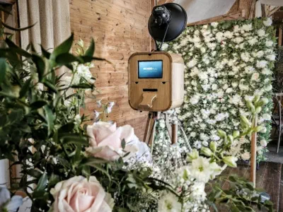 A vintage-style wooden photo booth stands on a tripod in front of a flower-covered backdrop. The surroundings are adorned with lush green leaves and various types of pastel-colored flowers, creating a cozy and elegant atmosphere.