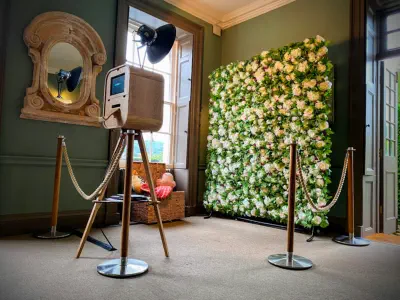 A photo booth setup in a green-painted room with an ornate mirror on the wall. The luxury photo booth hire features a camera on a stand, pointed towards a large backdrop adorned with white and green flowers, bordered by velvet ropes with brass stanchions.