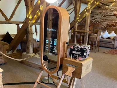 A wooden frame selfie mirror photo booth is set up indoors, adorned with fairy lights on the wooden beams in the background. Beside it is a small wooden box with a sign that reads, "Best Wedding Ever!" The rustic setting, reminiscent of Clock Barn photo b