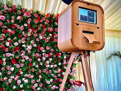 A vintage-style wooden photo booth stands on tripod legs in front of a vibrant flower wall composed of red, pink, and white roses. The booth features a small screen displaying images and a slot for photo printouts. A white, draped ceiling is visible above