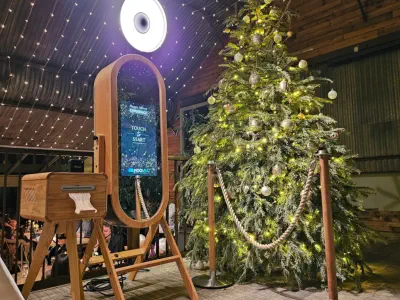 A wooden photo booth with a touch screen and an attached printer is set up beside a decorated Christmas tree under a canopy of string lights. The tree is adorned with white and yellow lights and ornaments. The scene is festive and inviting.
