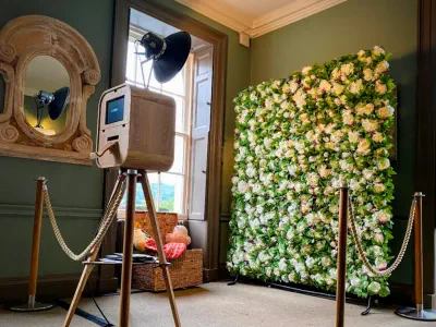 A photo booth setup in a stylish room, featuring a wooden camera on a tripod, a flash, and a backdrop wall adorned with white and green flowers. The booth is roped off with gold stanchions, and there's a wicker basket with props nearby. A large mirror han