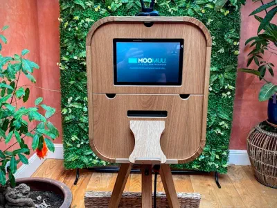 A wooden photo booth sits on a tripod in front of a lush green foliage backdrop. The screen on the booth displays "Moo Moo Photo Experience." Two potted plants flank the setup, adding a vibrant touch to the setting.