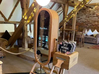 A rustic photo booth setup features an oval-shaped, full-length mirror with a light ring on top. The booth is made from wood and equipped with a camera. Nearby, a sign reads "Best Wedding Ever," with wooden beams and cozy seating in the background.