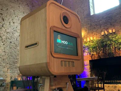 LA large, wooden, vintage-style luxury photo booth stands in a rustic stone-walled room. The photo booth has a rectangular screen displaying a logo and text. A circular camera lens is positioned above the screen. The room, perfect for luxury photo booth h