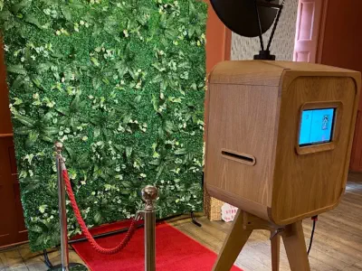 A photo booth setup features a modern wooden photo box with a digital screen facing a red carpet leading to a backdrop covered in green foliage. The scene is cordoned off with red ropes on silver stanchions, creating an elegant and inviting atmosphere.