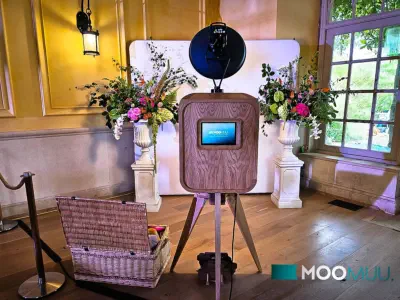 A wooden photo booth with a camera at the top and a screen displaying a tropical-themed image below stands in an indoor setting. In the background, a person is seated at a table, and another person is standing nearby. The atmosphere suggests a casual or s
