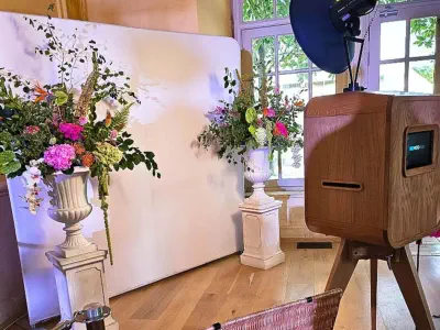 A wooden photo booth on a tripod stands in a lush indoor garden with various green plants and foliage. The booth has a small screen and a slot. The environment is well-lit with soft, natural light accentuating the greenery. A satellite dish is on top of t