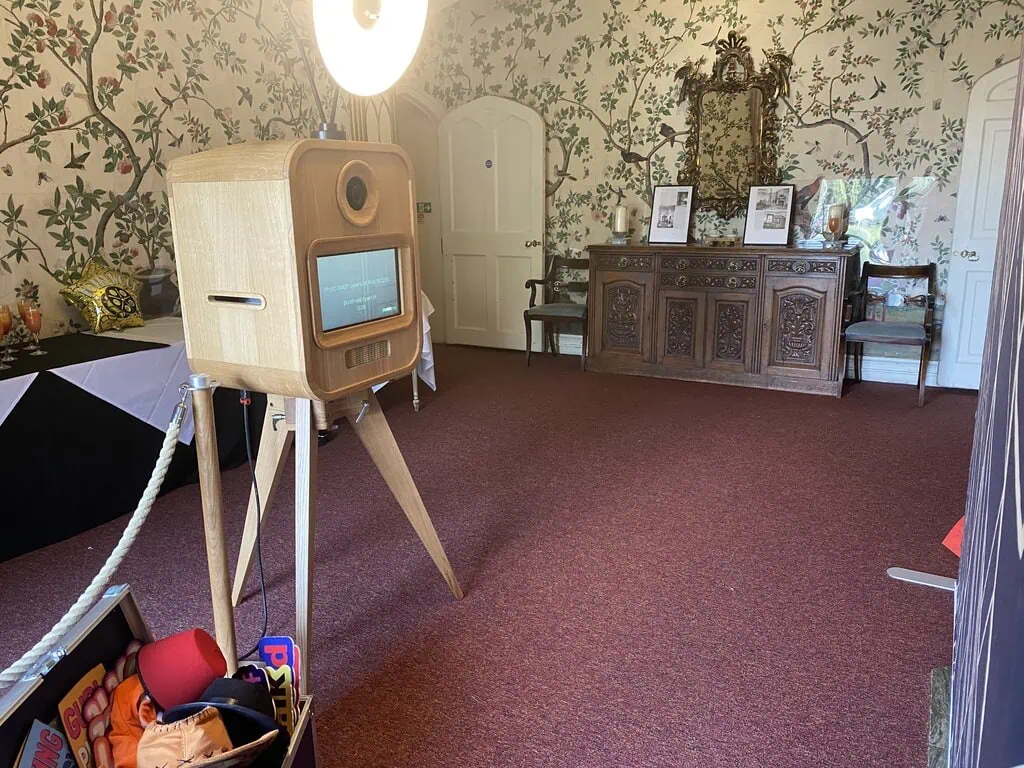 A wooden photo booth with a ring light stands in a carpeted room with floral wallpaper. An ornate sideboard with framed photos and a mirror lines the wall. Premium photo booth hire props are visible in a nearby basket.