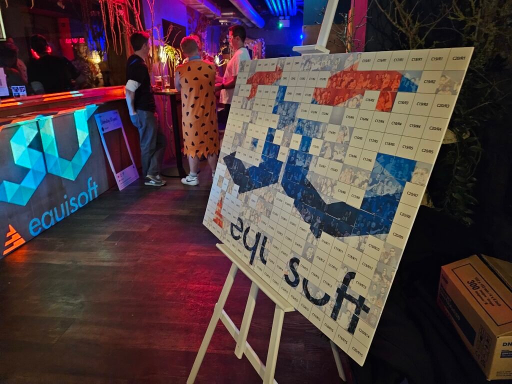 A mosaic board with coloured squares spelling "equisoft" stands on an easel at a dimly lit event, where guests socialise near the bar featuring an illuminated equisoft sign and premium photo booth hire.