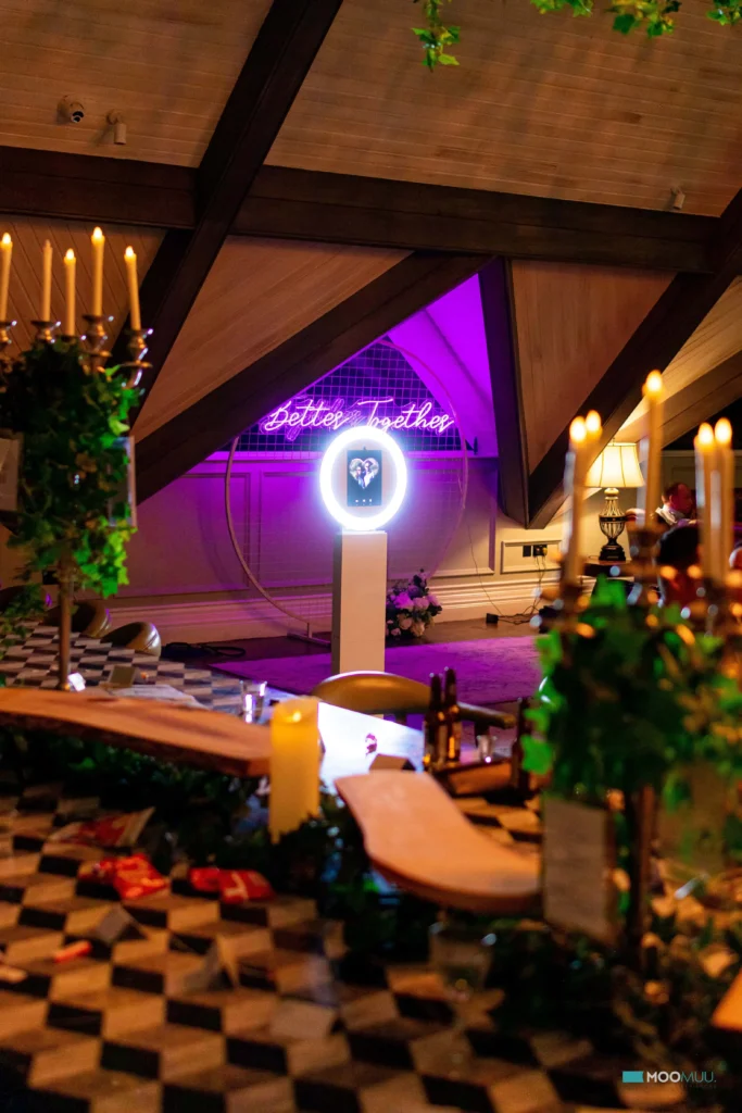 A warmly lit indoor event space features a neon “Better Together” sign on a purple-lit wall, premium photo booth hire with a circular booth, candles, lush greenery, and a chequered table set with snacks and decorative items.