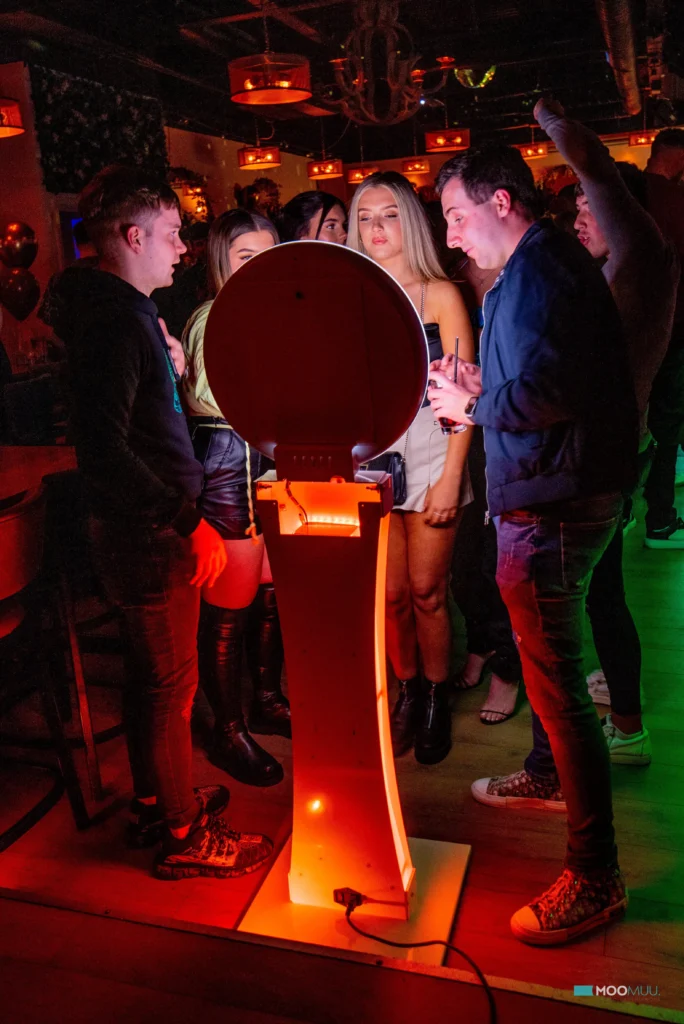 A group of people in a dimly lit bar gather round a photo booth, smiling and laughing as colourful lights illuminate their faces. Drinks and balloons set the scene for a fun corporate event experiential with sketch bot hire adding to the excitement.