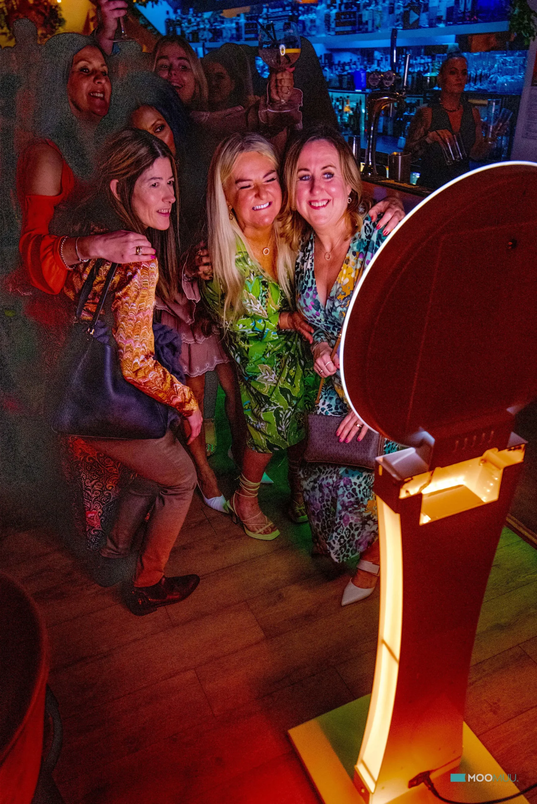 A group of smiling women in colourful outfits pose together for a photo in a lively, dimly lit indoor venue with warm lighting, visible bar elements, and premium photo booth hire adding a fun touch to the background.