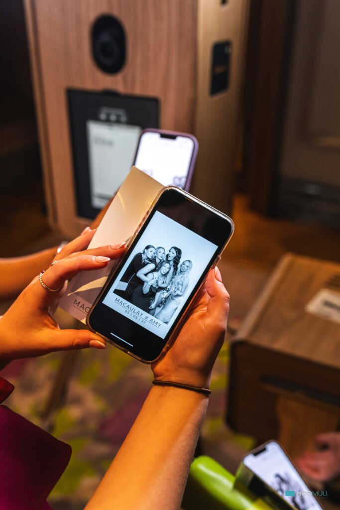 A person holds a mobile phone displaying a black-and-white photo of four smiling people posing closely together, likely at a corporate event experiential. Another hand and phone are visible in the background, and the setting appears to be indoors.