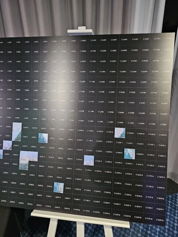 A large display board with small black squares labelled with codes, some featuring clouds or sky scenes. Perfect for a corporate event experiential, the board is set on an easel above a blue patterned carpet.