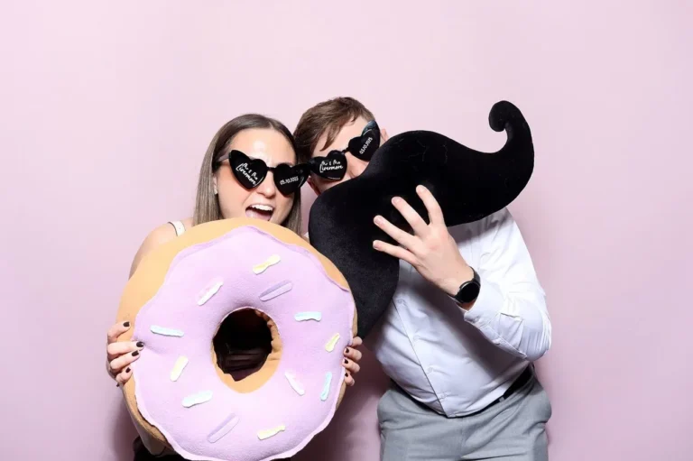 a couple standing and holding a giant donut and moustache with a pink backdrop