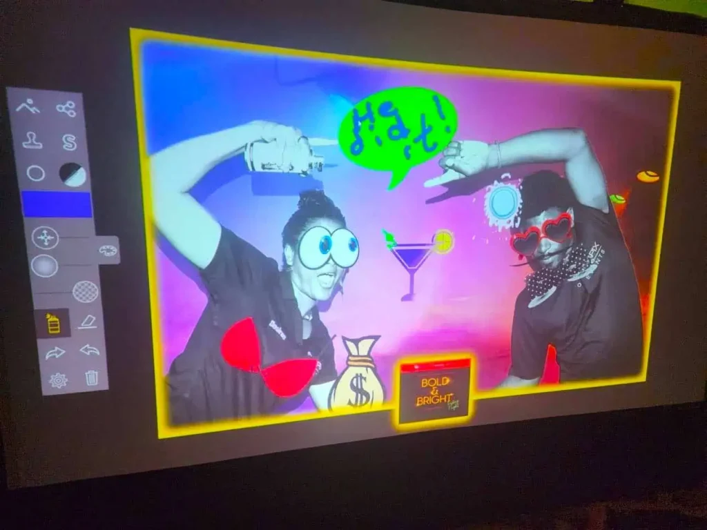 Two people pose playfully in a colourful, digitally-altered photo with cartoon sunglasses, speech bubbles, cocktails, and a "Bold & Bright" sign—perfect inspiration for a corporate event experiential or graffiti wall hire activation.
