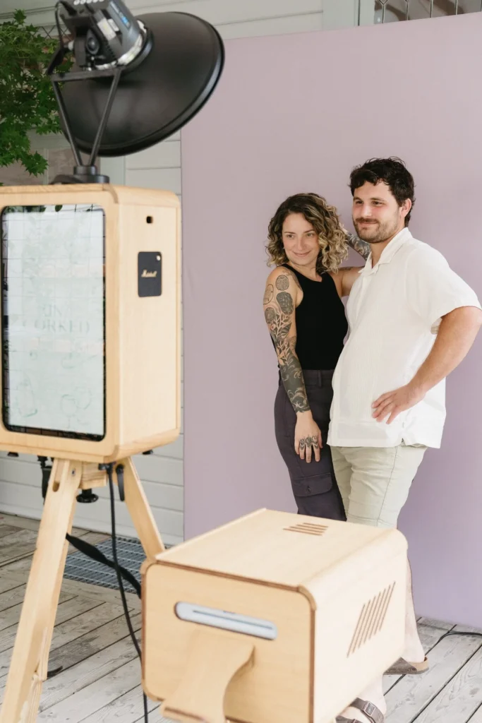 A smiling couple pose together in front of a light purple backdrop, standing close with arms round each other, next to a wooden photo booth set-up with studio lighting—perfect for a corporate event experiential or graffiti wall hire.