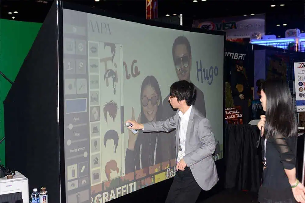 A man uses a digital pen to draw on a large interactive screen displaying faces and drawing tools, whilst a woman watches beside him at an indoor corporate event experiential or exhibition, showcasing features similar to premium photo booth hire.