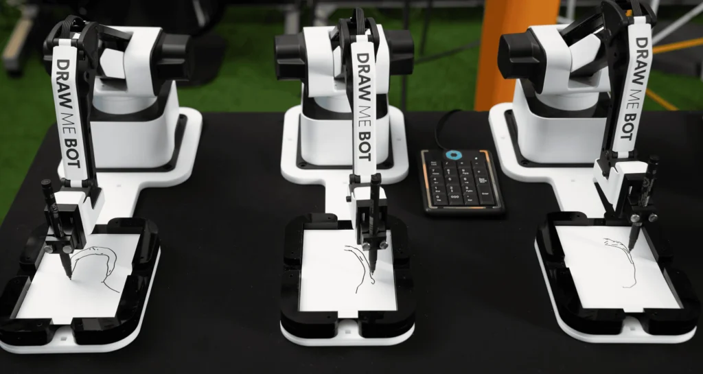 Three robotic arms labelled "DRAW ME BOT" are sketching unique outlines on paper, accompanied by a small keypad beside the middle robot, all arranged on a black table—perfect for adding a creative twist to your corporate event experiential.