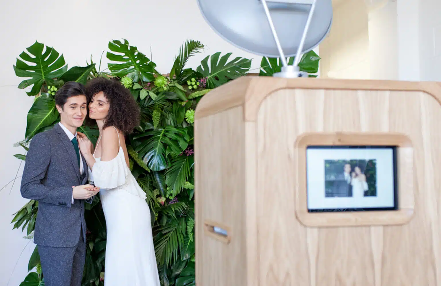 Wedding couple sharing a kiss beside the Luxury Oak Booth and a green foliage backdrop.