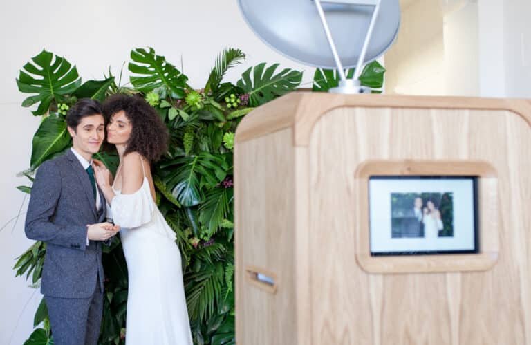 A couple pose in front of a leafy green backdrop, with one person kissing the other on the cheek. A retro wooden photo booth with their image on the screen is visible in the foreground.