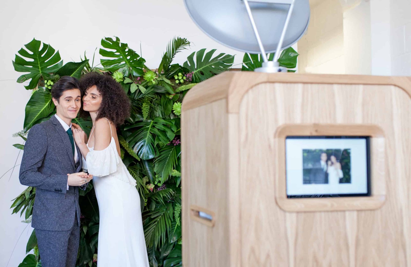 A couple pose in front of a leafy green backdrop, with one person kissing the other on the cheek. A retro wooden photo booth with their image on the screen is visible in the foreground.