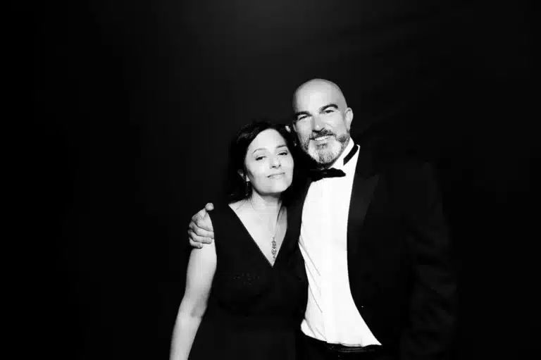 A man in a dinner jacket and a woman in a sleeveless dress stand together, smiling with the man's arm around her shoulder, capturing the joy of celebrations against a plain dark background. The image is in black and white.