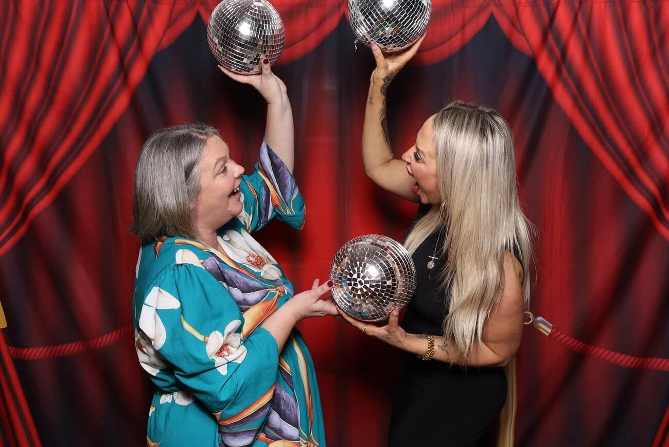 Two women stand in front of a red curtain backdrop, smiling and holding shiny disco balls above their heads, exuding the joyful spirit of celebrations.