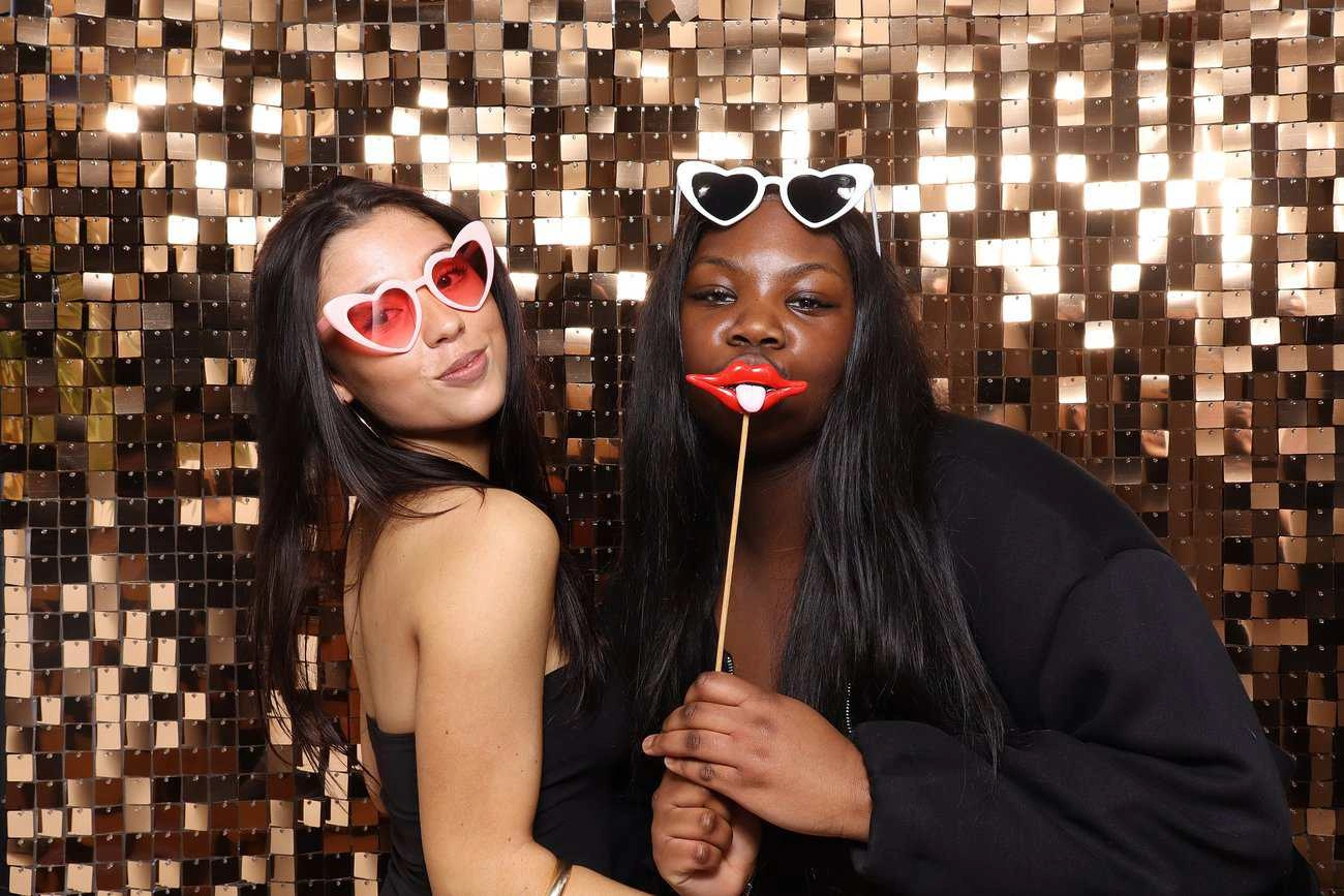 Two women pose in front of a sparkly gold sequin backdrop, celebrating with heart-shaped sunglasses. One holds playful red lips on a stick. Both smile in dark outfits, capturing the joy of celebrations in this fun photo booth moment.