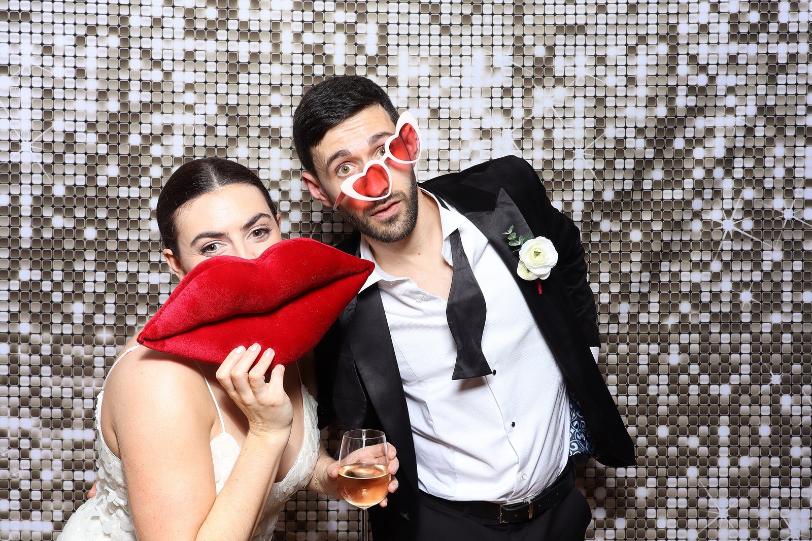 A woman in a white dress holds large red lips props, and a man in a suit with an untied bow tie and heart-shaped glasses holds a drink. Both pose playfully in front of a sparkling graffiti wall hire photo booth backdrop.