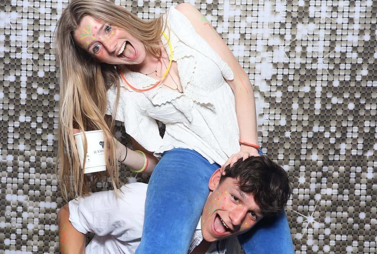 A young woman in a white top sits on a young man's shoulders, both smiling and laughing during celebrations. She holds a cup, their faces splashed with colourful paint, against a shiny, tiled background.