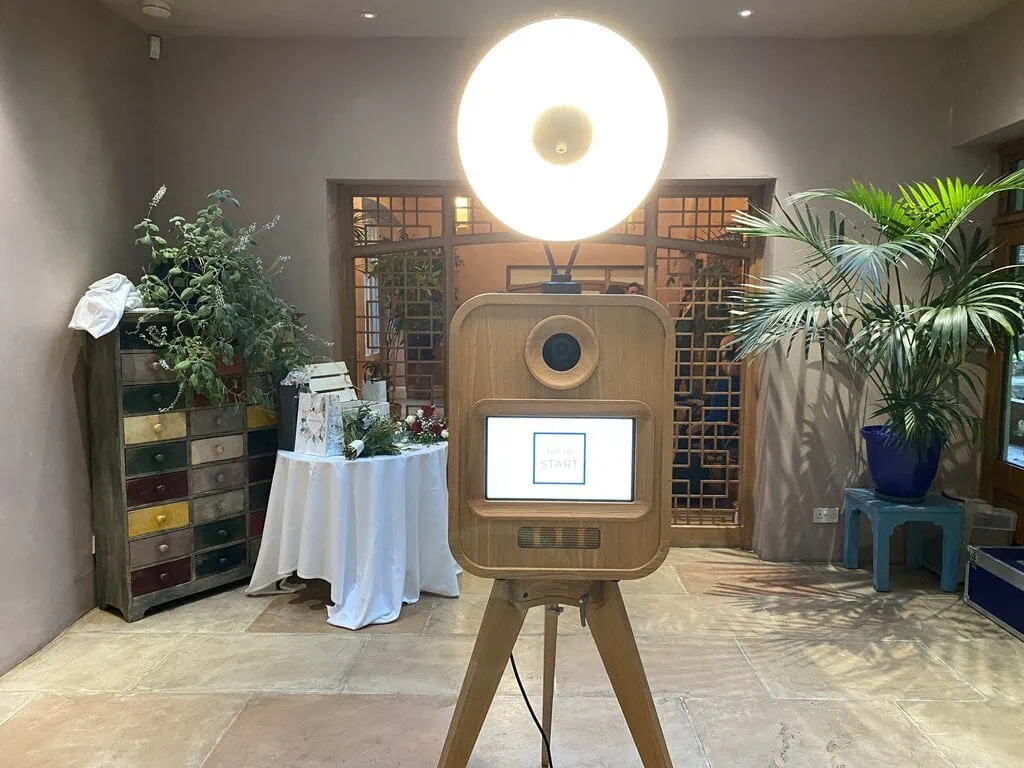 A vintage-style photo booth with a bright circular light stands in a room with potted plants, decor items on a table, and a colourful drawer cabinet. Perfect for corporate event experiential moments or premium photo booth hire. The screen displays "Touch to Start.