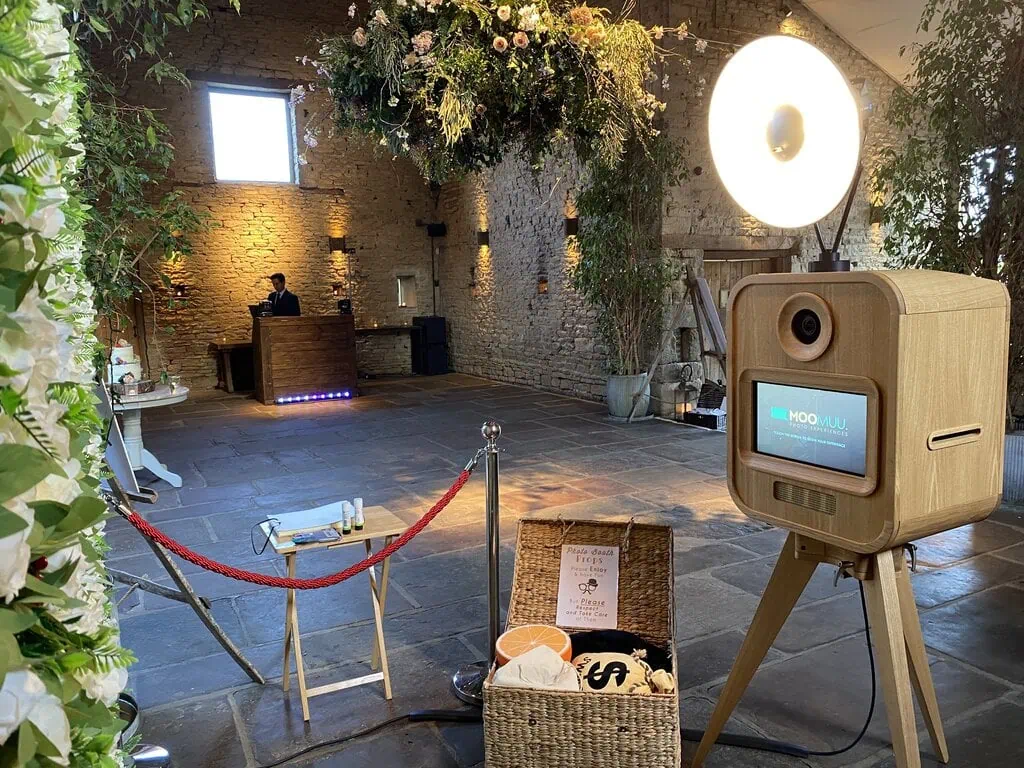 A premium photo booth hire with a ring light stands in a rustic venue with exposed brick walls, floral decorations, and a roped-off area. A person sits at a desk in the distance, and various props are placed on the floor nearby.