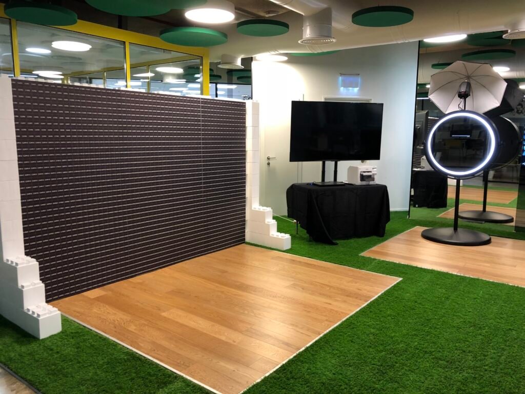 A modern photo booth setup for a corporate event experiential features a blank LEGO wall, a TV, ring light with umbrella, and printer on a black table, all arranged on artificial grass flooring in a brightly lit indoor space.