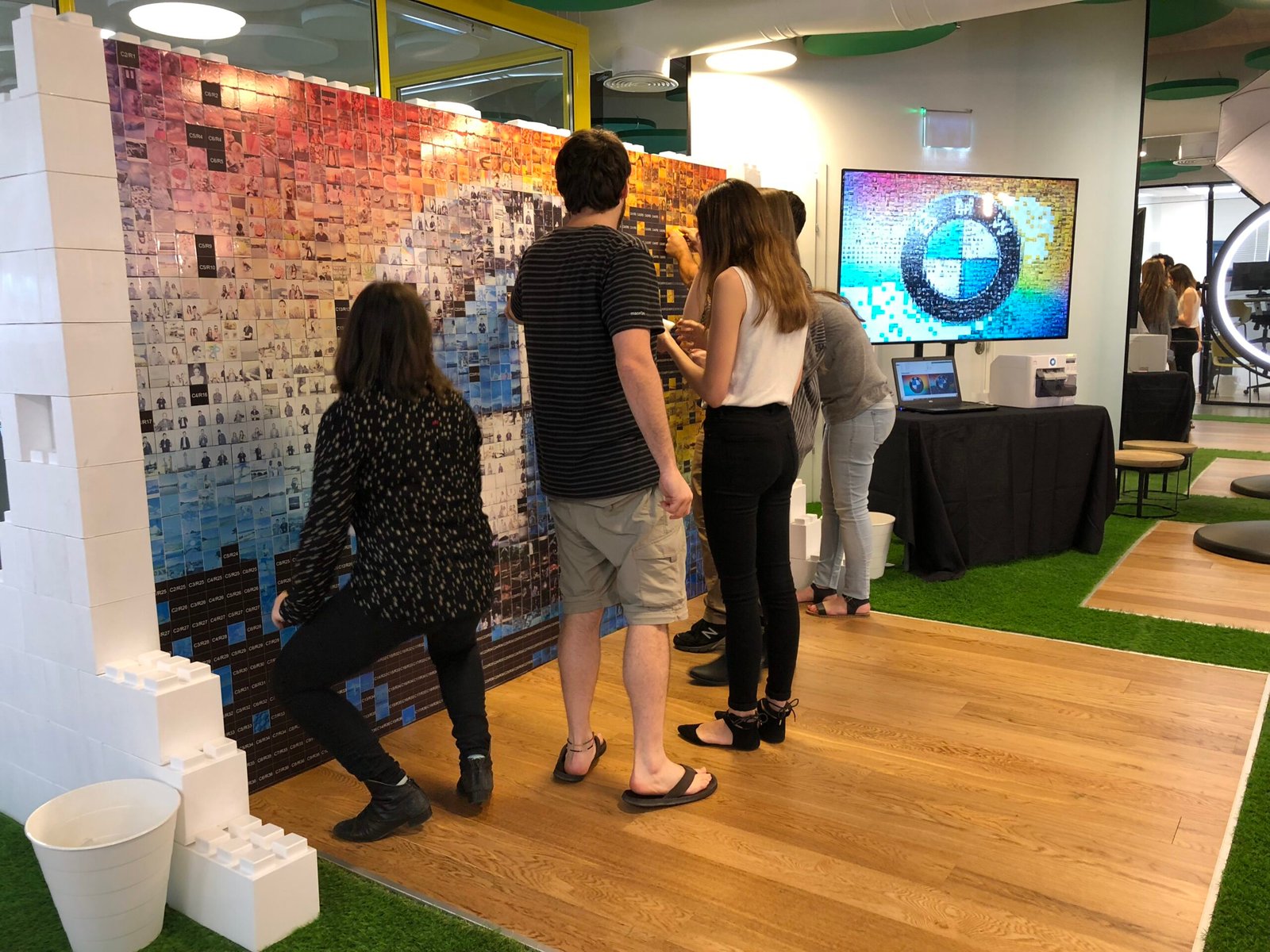 Four people stand in front of a colourful photo mosaic wall, adding tiles to form the BMW logo. Highlighting a corporate event experiential atmosphere, a screen displays the same logo in a modern office space.