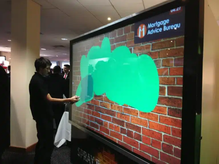 A person uses a large interactive touchscreen to virtually paint green shapes over a brick wall image. The screen displays the Mortgage Advice Bureau logo in the top right corner.