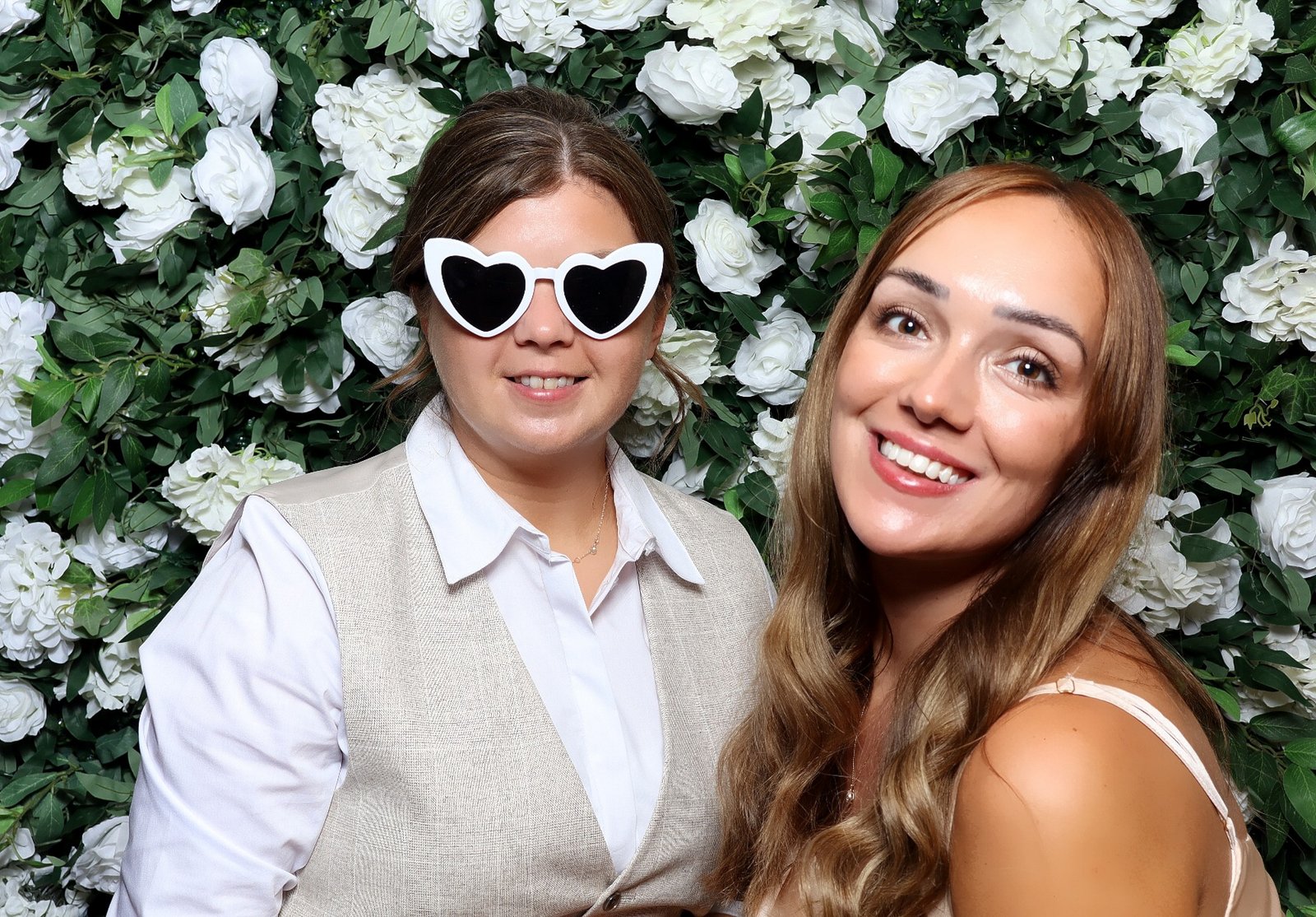 Two guests smiling in a MooMuu colour portrait against a white flower and foliage backdrop