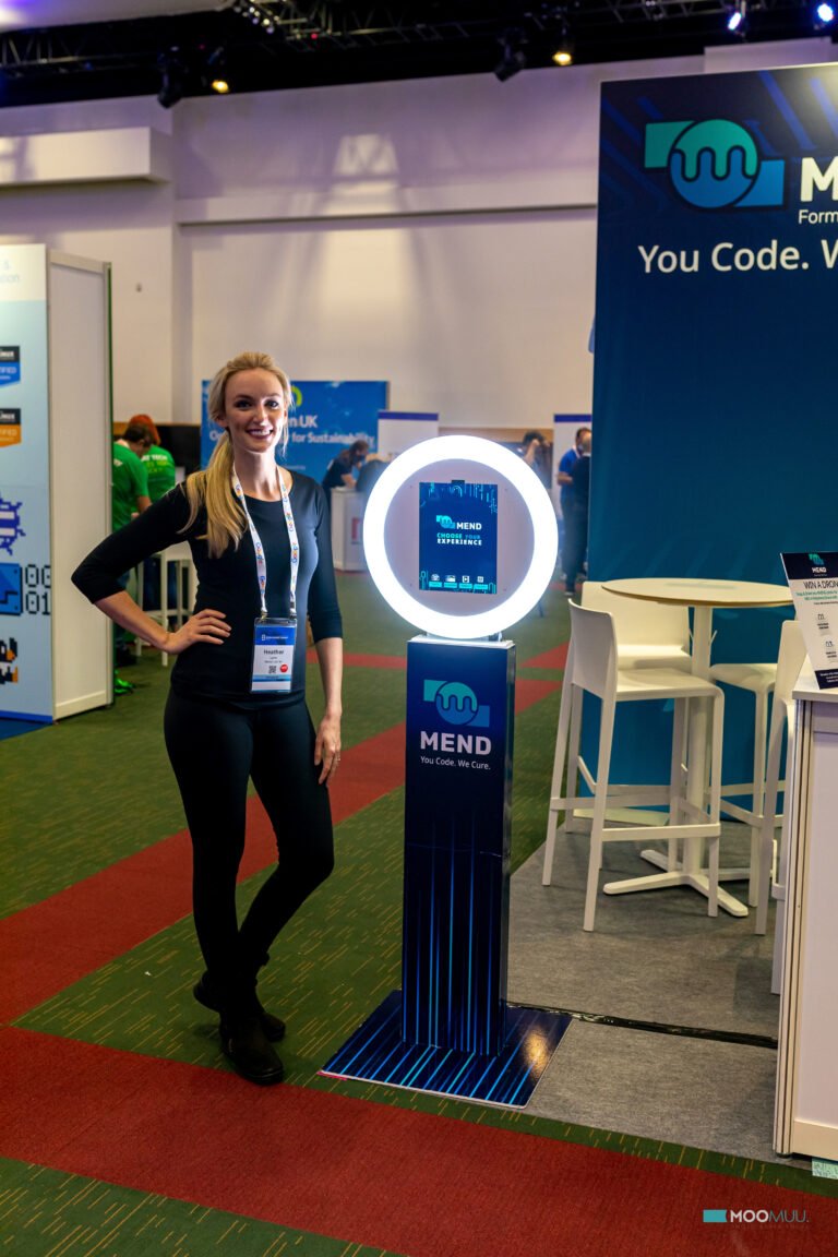 A woman in black stands next to a ring light photo booth at a Mend software stand during a tech event. The stand displays the Mend logo and slogan, “You Code. We Secure.” There are chairs and tables in the background.