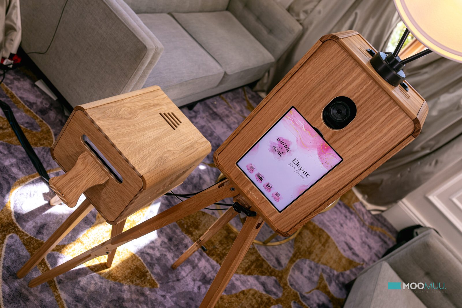 A wooden photo booth with a touch screen and camera lens stands on a tripod, next to a matching wooden box with a slot, in a modern living room with grey furniture and patterned carpet.