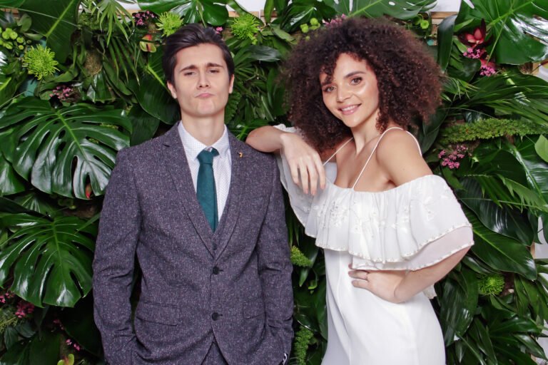 Two people pose in front of a lush wall of green leaves. One wears a grey suit and teal tie, while the other wears a white off-the-shoulder dress and rests an arm on their companion’s shoulder.